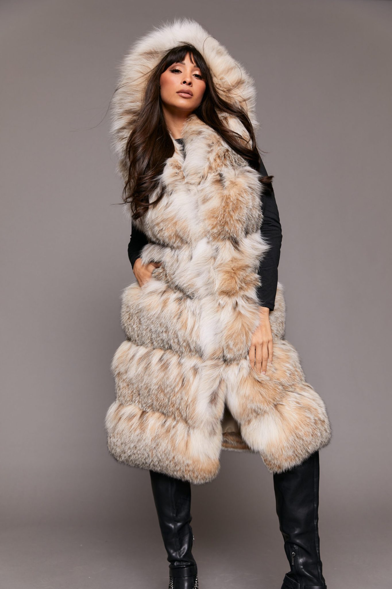 Frontal view of a woman wearing a fur vest with the hood up standing in front of a grey background