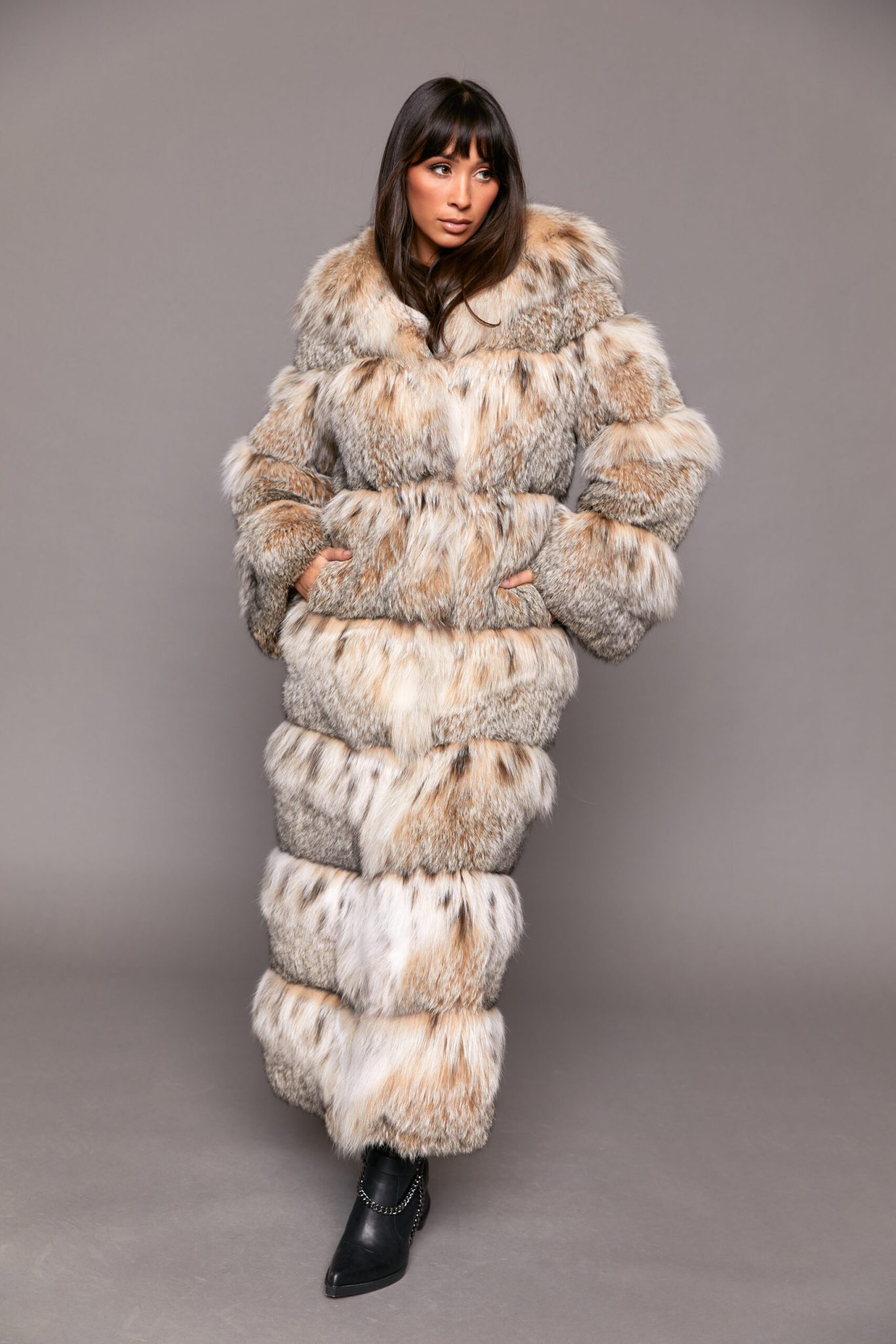Frontal view of a woman wearing a full length fur coat with her arms on her hips standing in front of a grey background