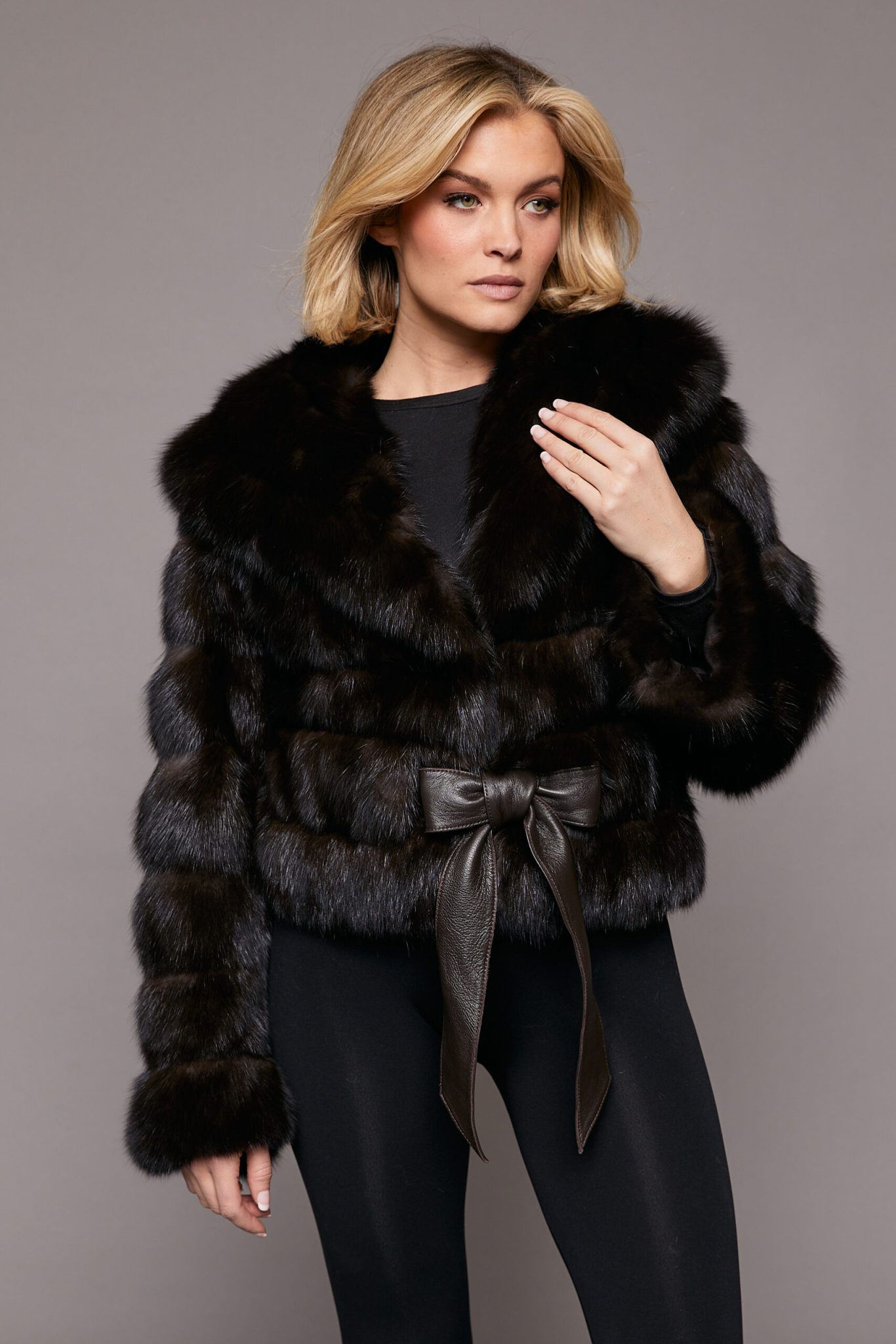 Frontal view of a woman wearing a sable jacket with her left hand raised and touching the collar, standing in front of a grey background
