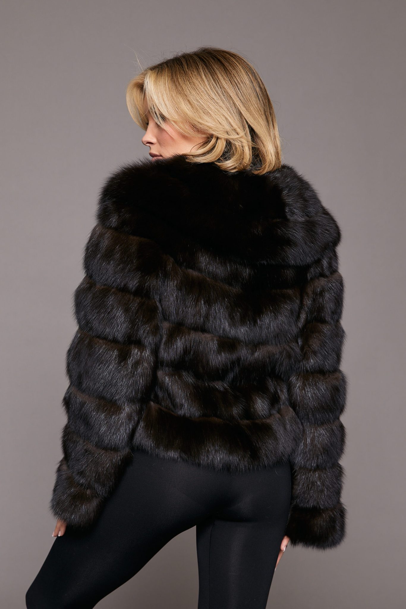 Rear view of a woman wearing a sable jacket standing towards a grey background, her head is sloghtly turned to the left