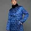 Frontal view of a woman in a reversible coat with the blue quilted shell side out, she has her left hand in the corresponding front pocket and is standing in front of a grey background