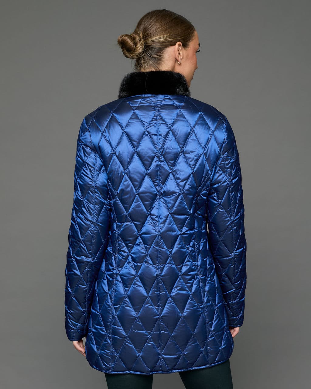 Reverse view of a woman in a reversible coat with the blue quilted shell side out, she both arms positioned straight down to her sides and is standing facing a grey background