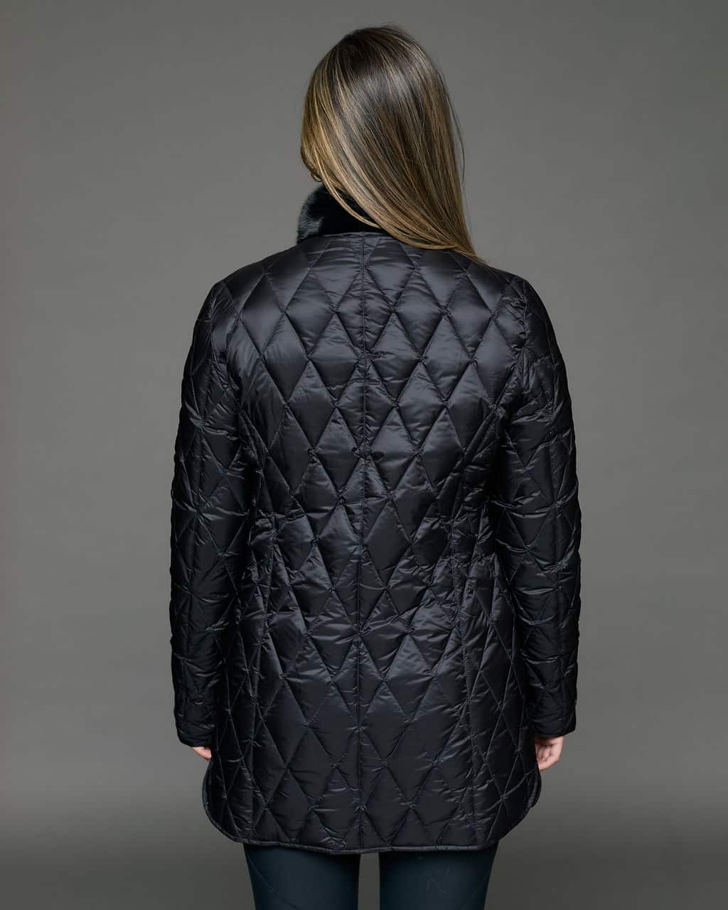 Rear view of a woman in a reversible coat with the black quilted shell side out, she has both arms at her sides and is facing a grey background