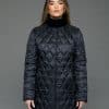 Frontal view of a woman in a reversible coat with the black quilted shell side out, she has both arms at her sides and is standing in front of a grey background