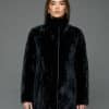 Frontal view of a woman with long brown hair in a reversible coat with the black fur side out, she has both arms at her sides and is standing in front of a grey background