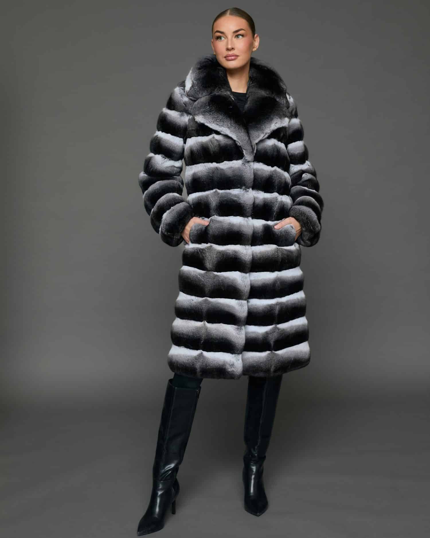 Frontal view of a woman in a natural empress chinchilla coat with her hands in the pockets standing in front of a grey background.