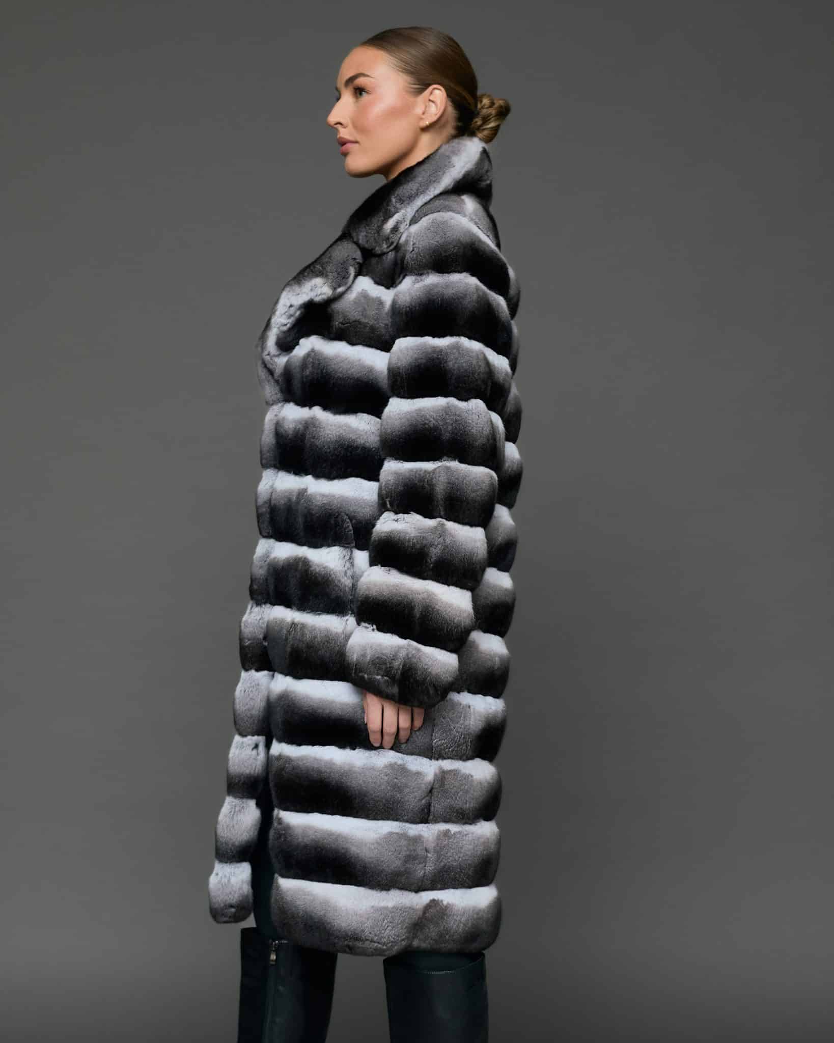 ECOMMWOMENS20255569 Profile shot of a woman in a natural empress chinchilla coat with her hands at her side standing in front of a grey background.