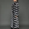 ECOMMWOMENS20255583 Profile view of a woman in a natural chinchilla full length coat standing against a grey background