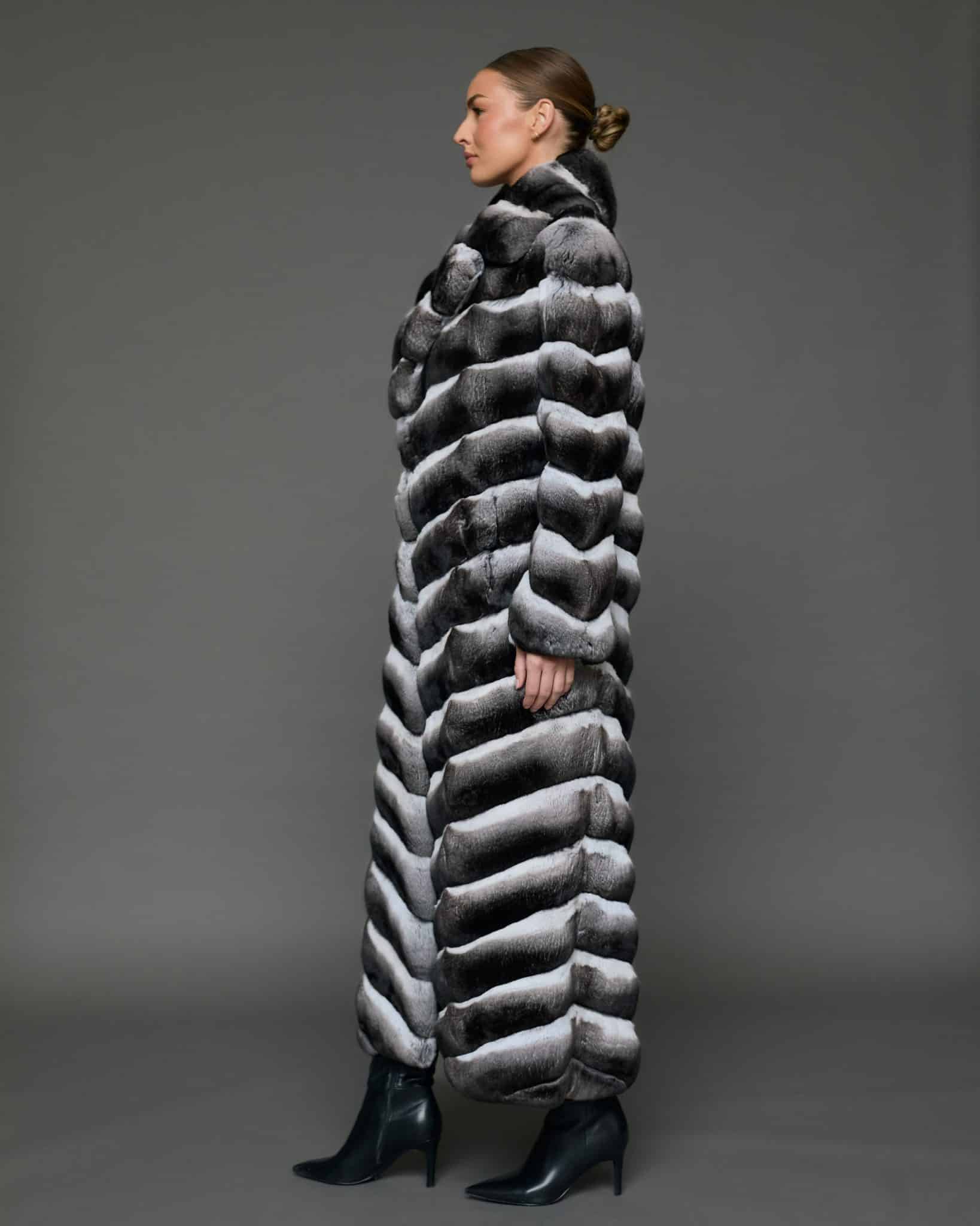 ECOMMWOMENS20255583 Profile view of a woman in a natural chinchilla full length coat standing against a grey background