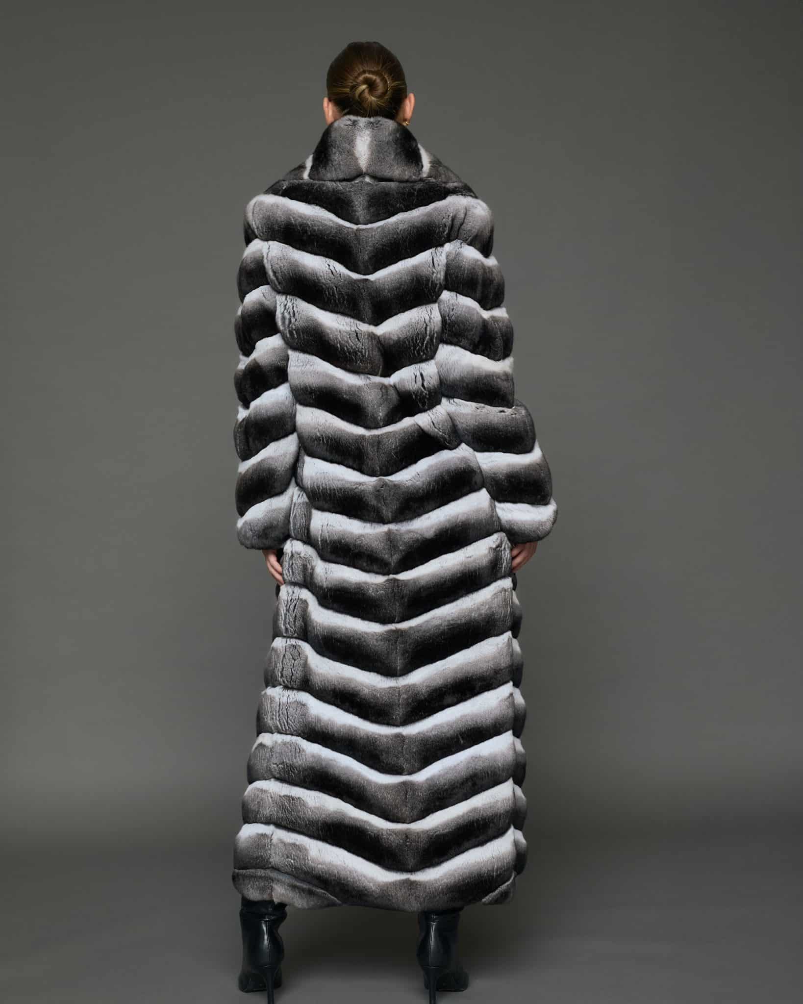 ECOMMWOMENS20255584 Rear view of a woman in a natural chinchilla full length coat, facing a grey background