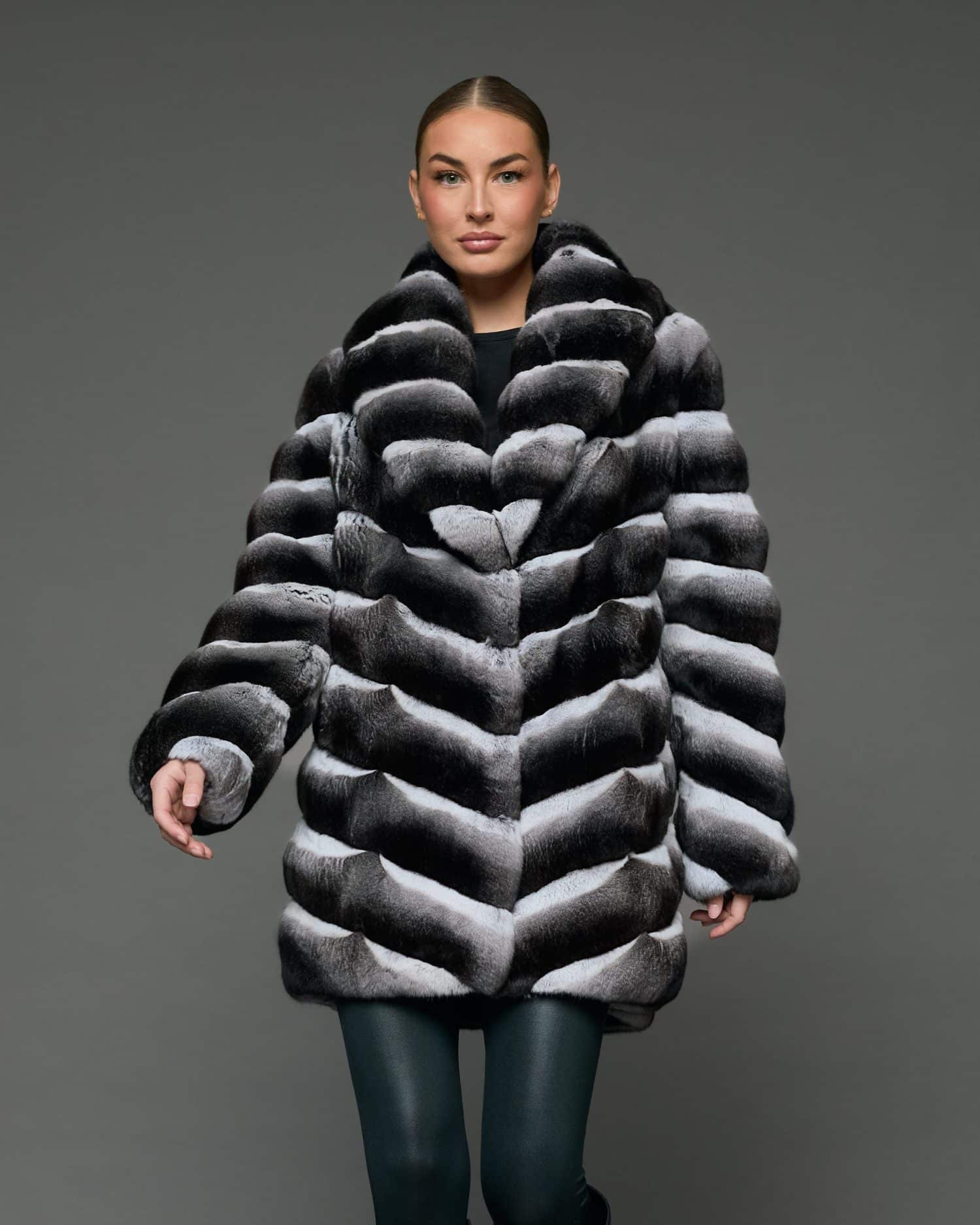 Frontal view of a woman in a stroller length chinchilla coat with a shawl collar standing in front of a grey background