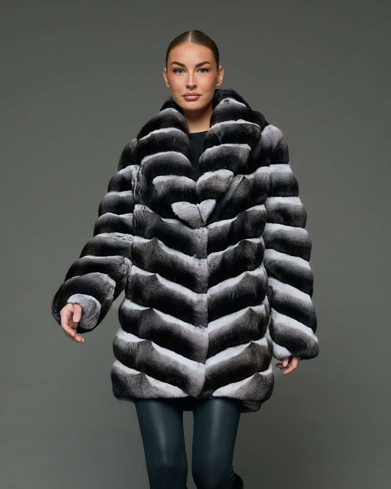 Frontal view of a woman in a stroller length chinchilla coat with a shawl collar standing in front of a grey background