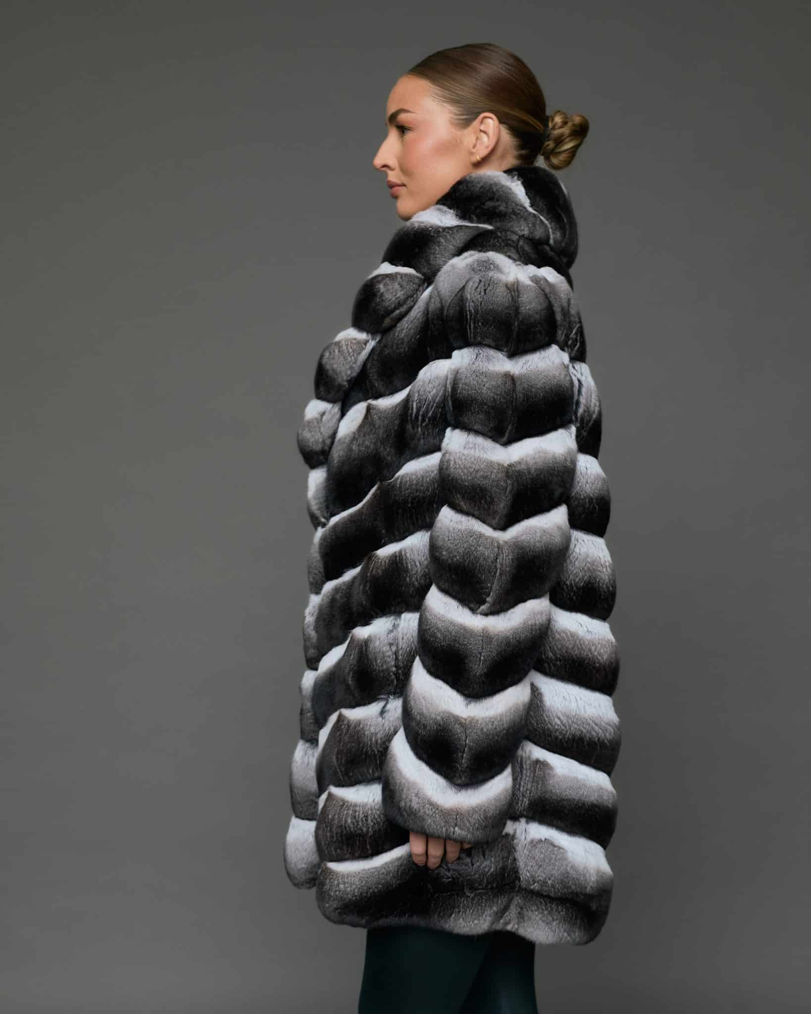 Profile view of a woman in a stroller length chinchilla coat with a shawl collar standing in front of a grey background