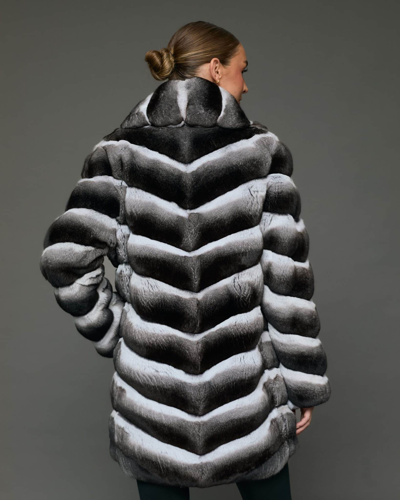 Rear view of a woman in a stroller length chinchilla coat with a shawl collar facing a grey background