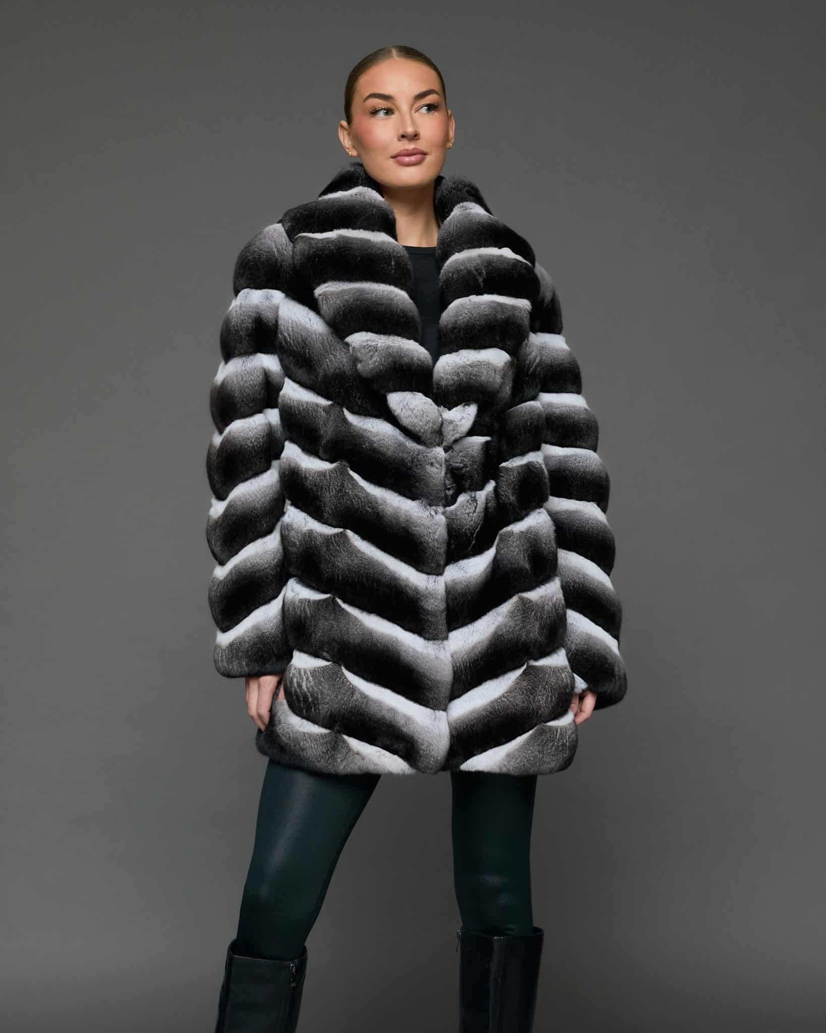 Frontal view of a woman in a stroller length chinchilla coat with a shawl collar standing in front of a grey background, the woman's eyes are looking off to stage right