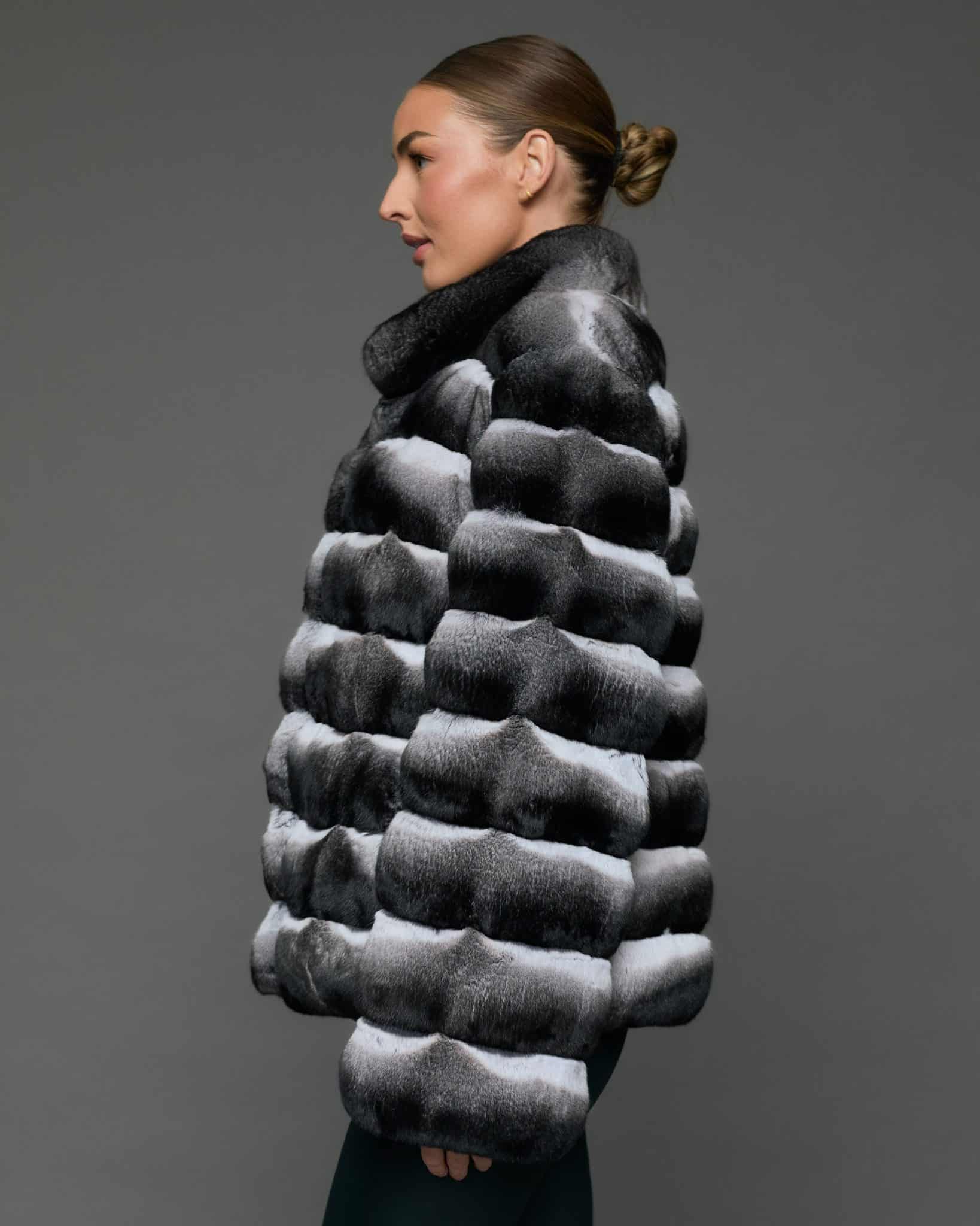 ECOMMWOMENS20255645 Profile view of a woman in a natural black and white chinchilla jacket with her arms at her sides standing in front of a grey background