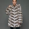 Frontal view of a woman in a natural beige chinchilla coat with the hood down standing in front of a grey background