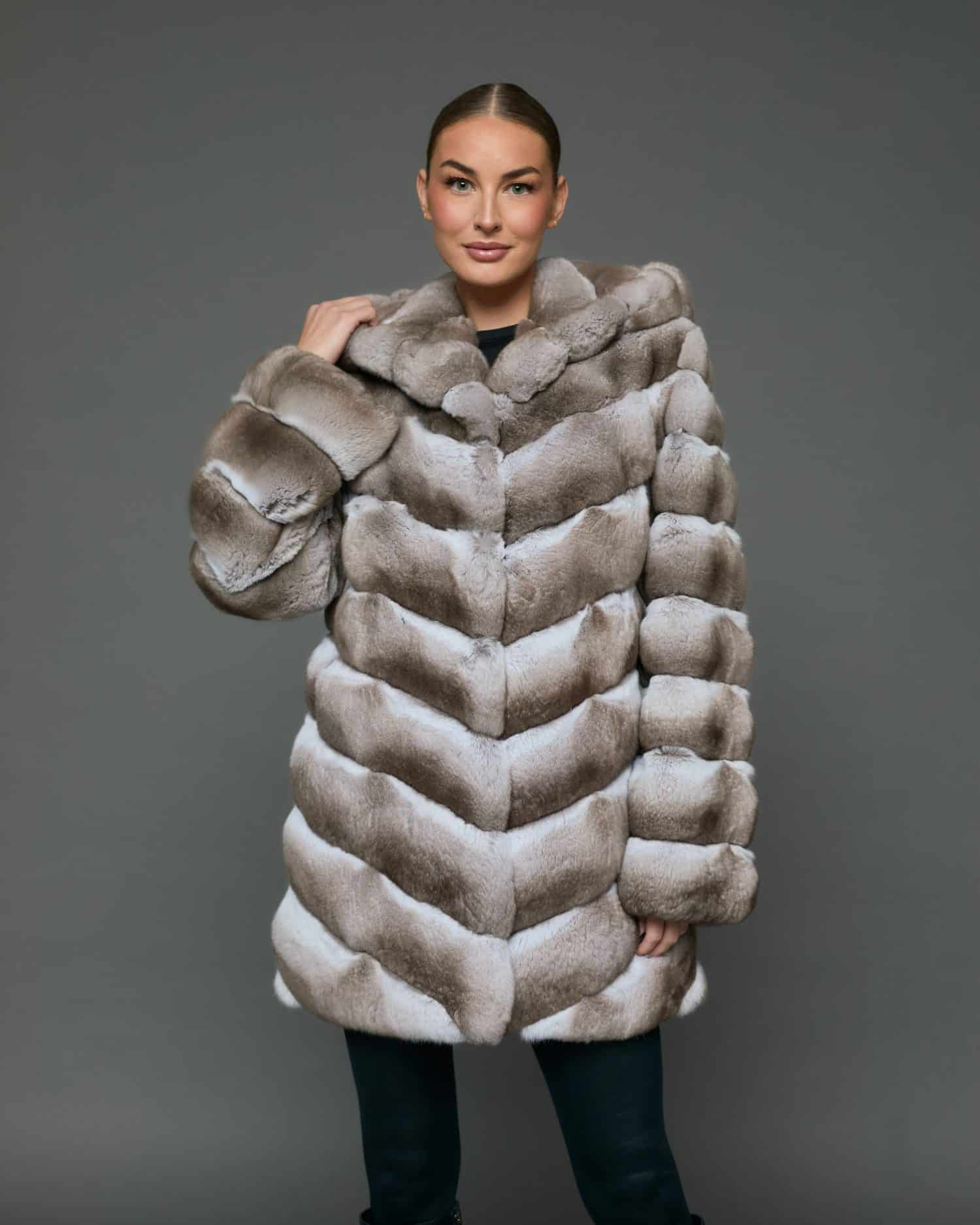 Frontal view of a woman in a natural beige chinchilla coat with the hood down standing in front of a grey background