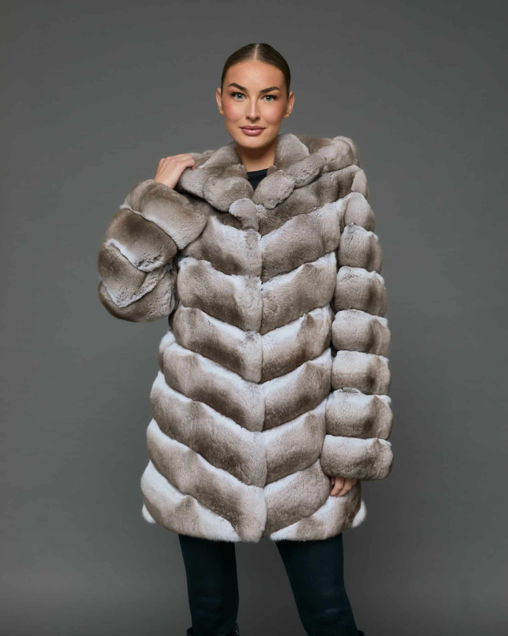 Frontal view of a woman in a natural beige chinchilla coat with the hood down standing in front of a grey background