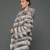 Side view of a woman in a natural beige chinchilla coat with the hood down standing in front of a grey background