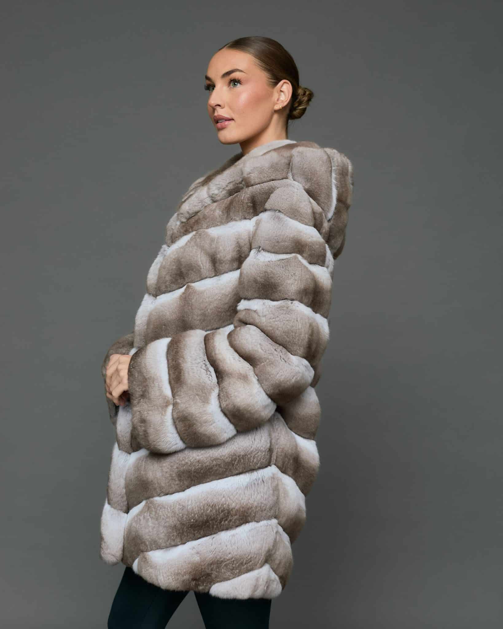 Side view of a woman in a natural beige chinchilla coat with the hood down standing in front of a grey background