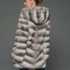 Rear view of a woman in a natural beige chinchilla coat with the hood down standing in front of a grey background