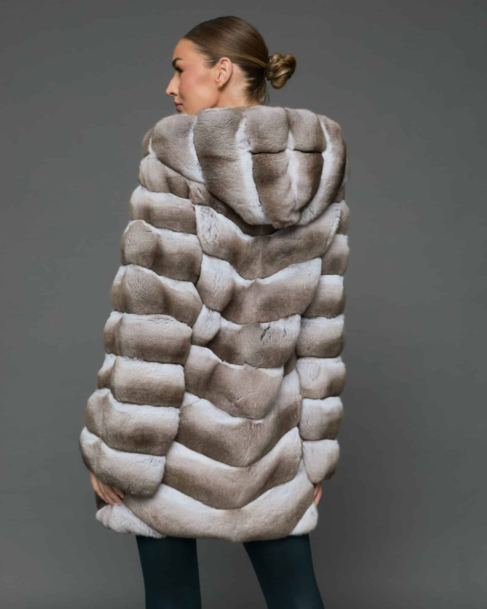 Rear view of a woman in a natural beige chinchilla coat with the hood down standing in front of a grey background
