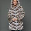 Frontal view of a woman in a natural beige chinchilla coat with the hood up standing in front of a grey background
