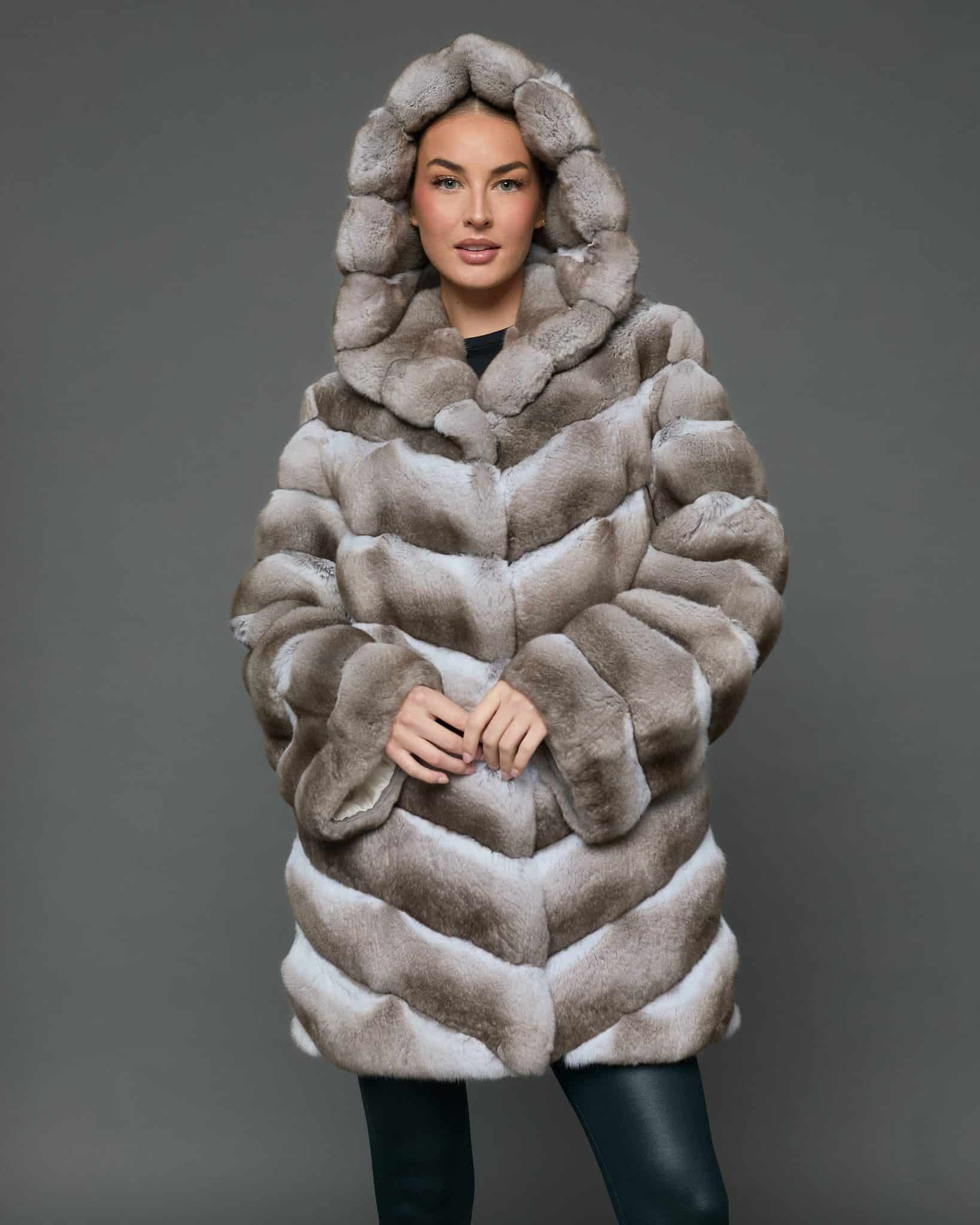 Frontal view of a woman in a natural beige chinchilla coat with the hood up standing in front of a grey background