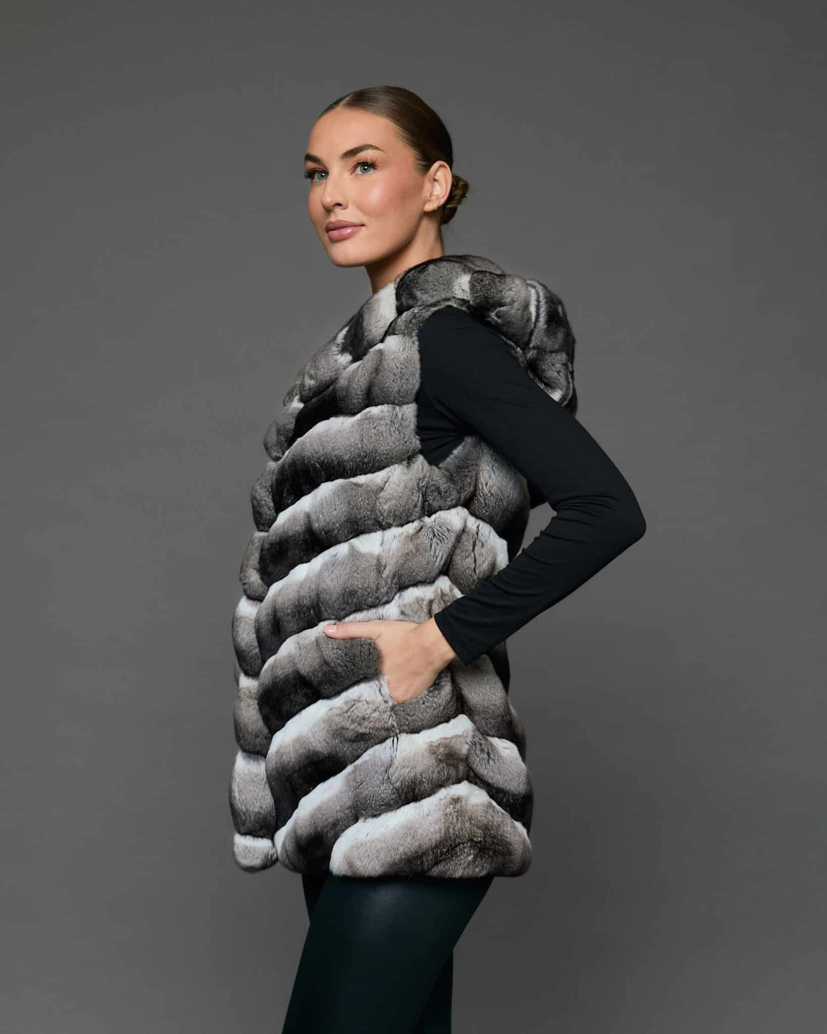Side view of a woman in a sleeveless chinchilla vest with her left hand in the vest pocket standing against a grey background