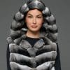 Close-up view of a woman in a sleeveless chinchilla vest with the hood up standing against a grey background