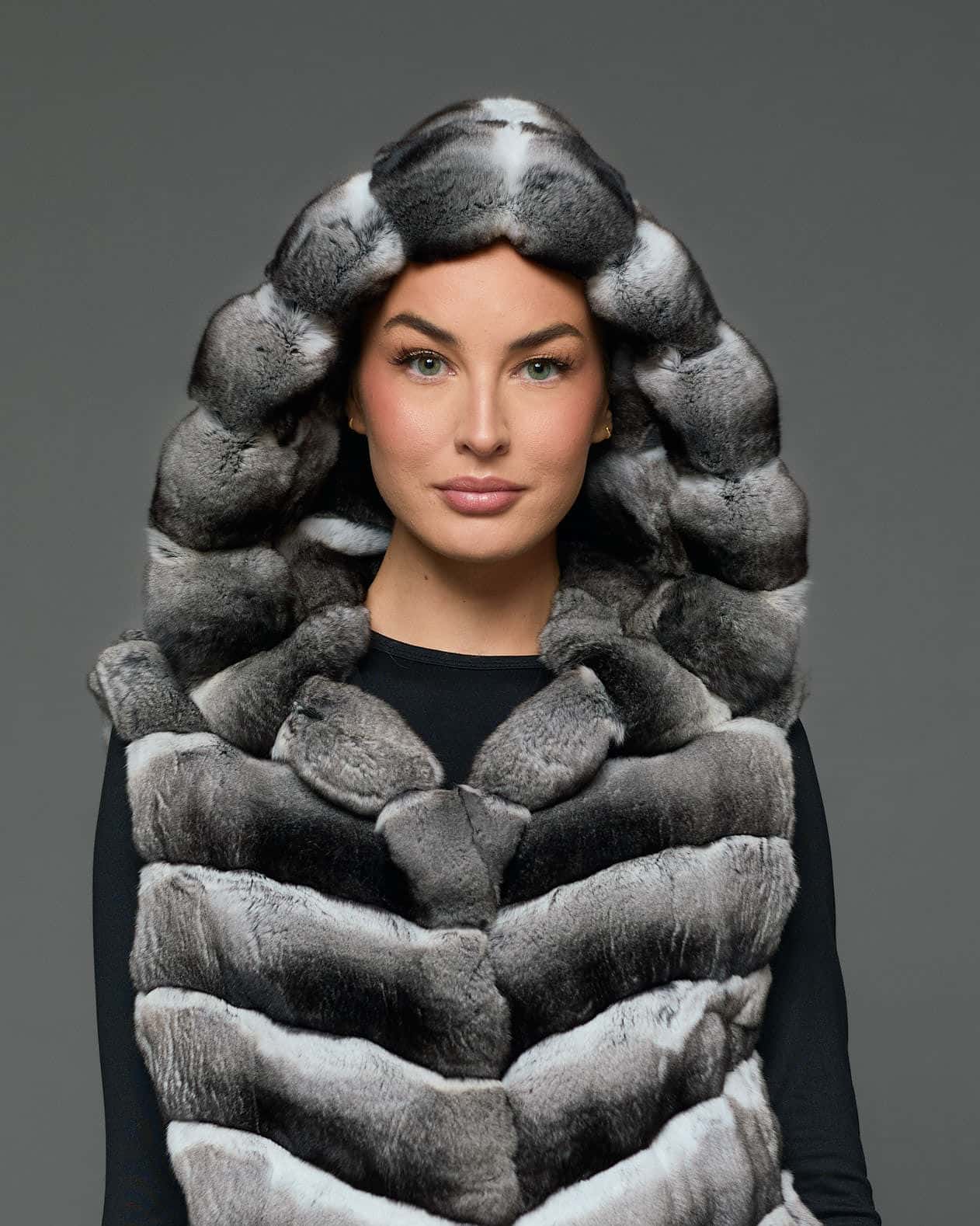 Close-up view of a woman in a sleeveless chinchilla vest with the hood up standing against a grey background