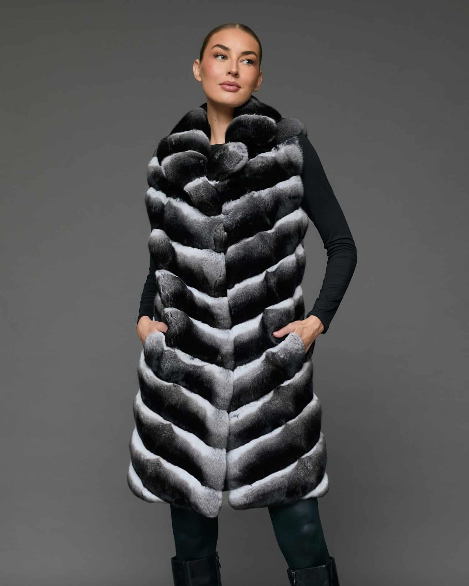 Front view of a woman in a knee-length black and white chinchilla vest standing in front of a grey background