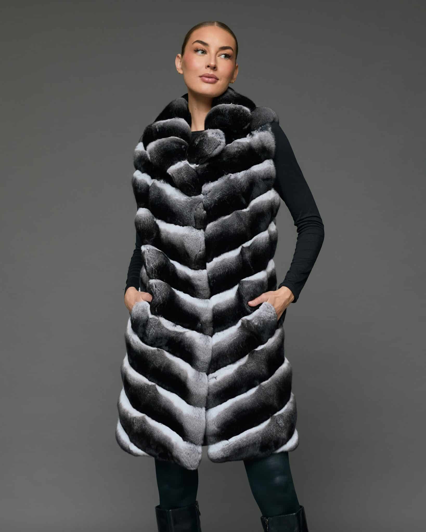 Front view of a woman in a knee-length black and white chinchilla vest standing in front of a grey background