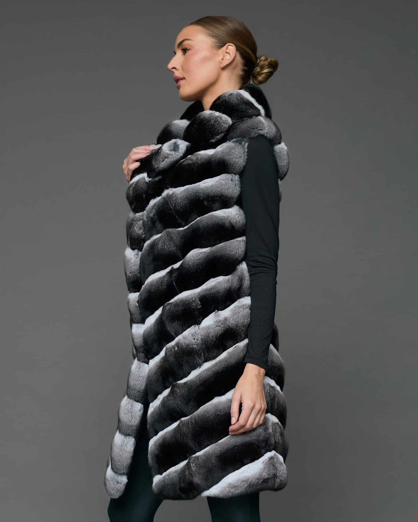 Side view of a woman in a knee-length black and white chinchilla vest standing in front of a grey background