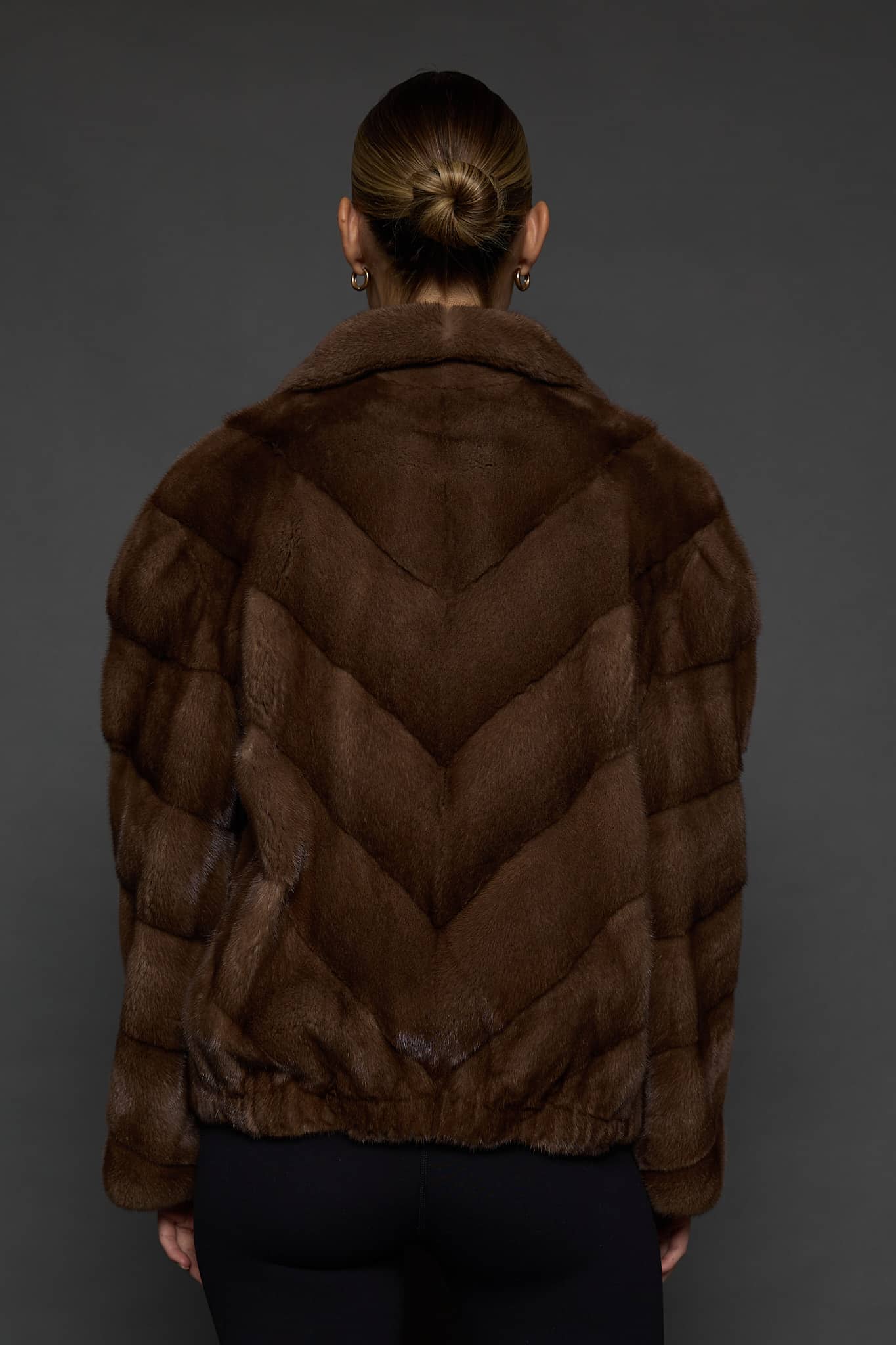 Rear view of a woman in a brown mink jacket facing a dark grey background.