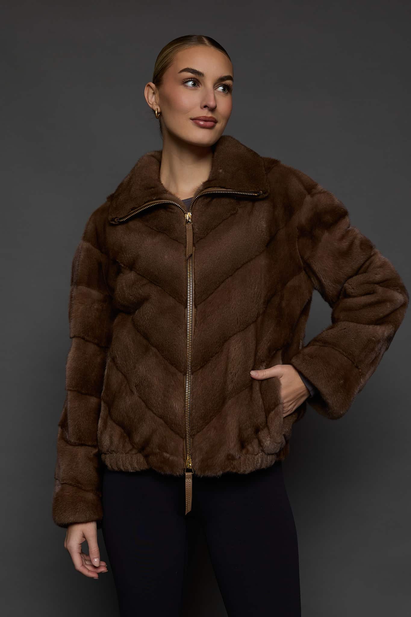 Frontal view of a woman in a brown mink jacket standing in front of a dark grey background.