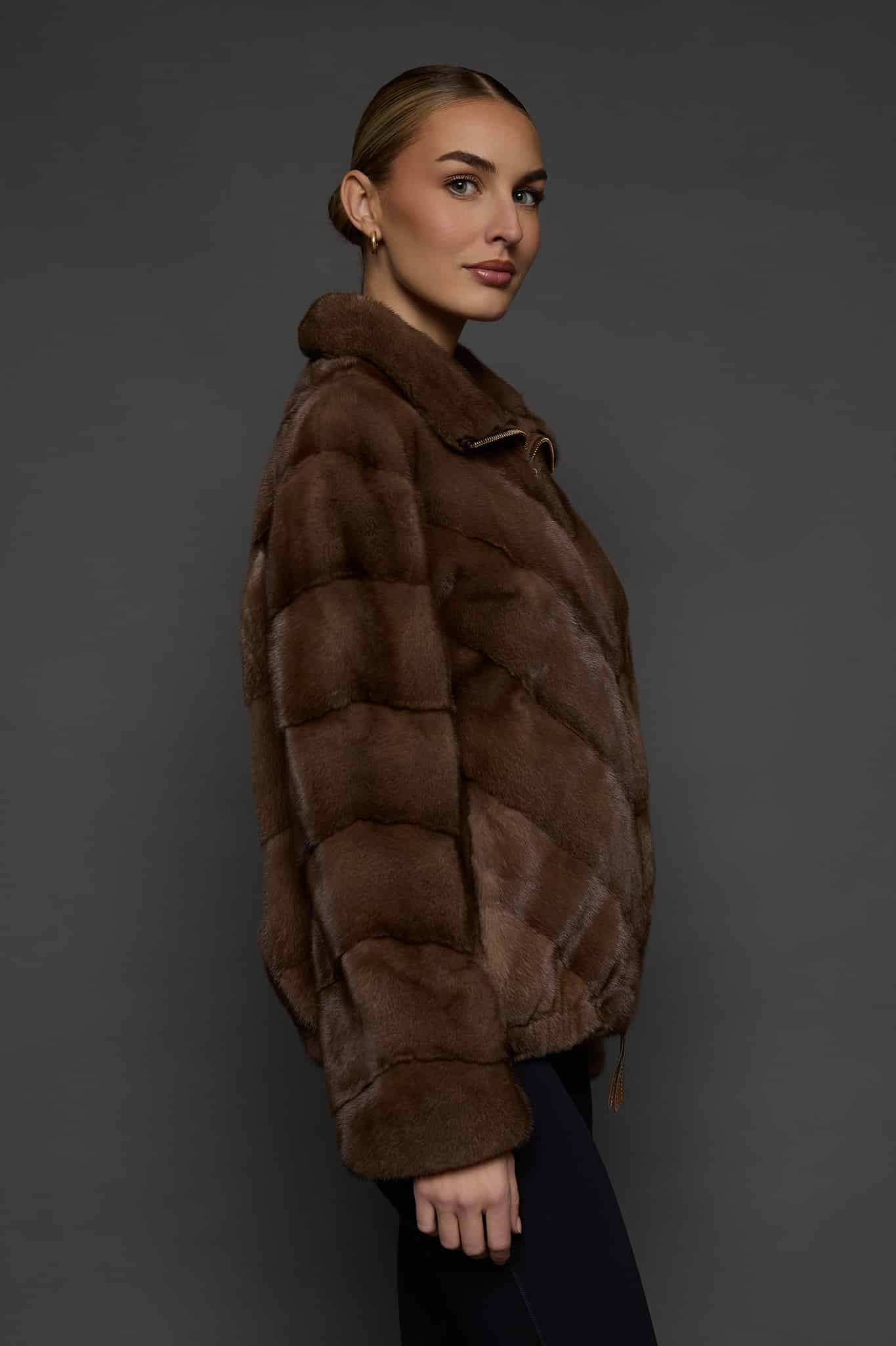 Profile view of a woman in a brown mink jacket standing in front of a dark grey background.