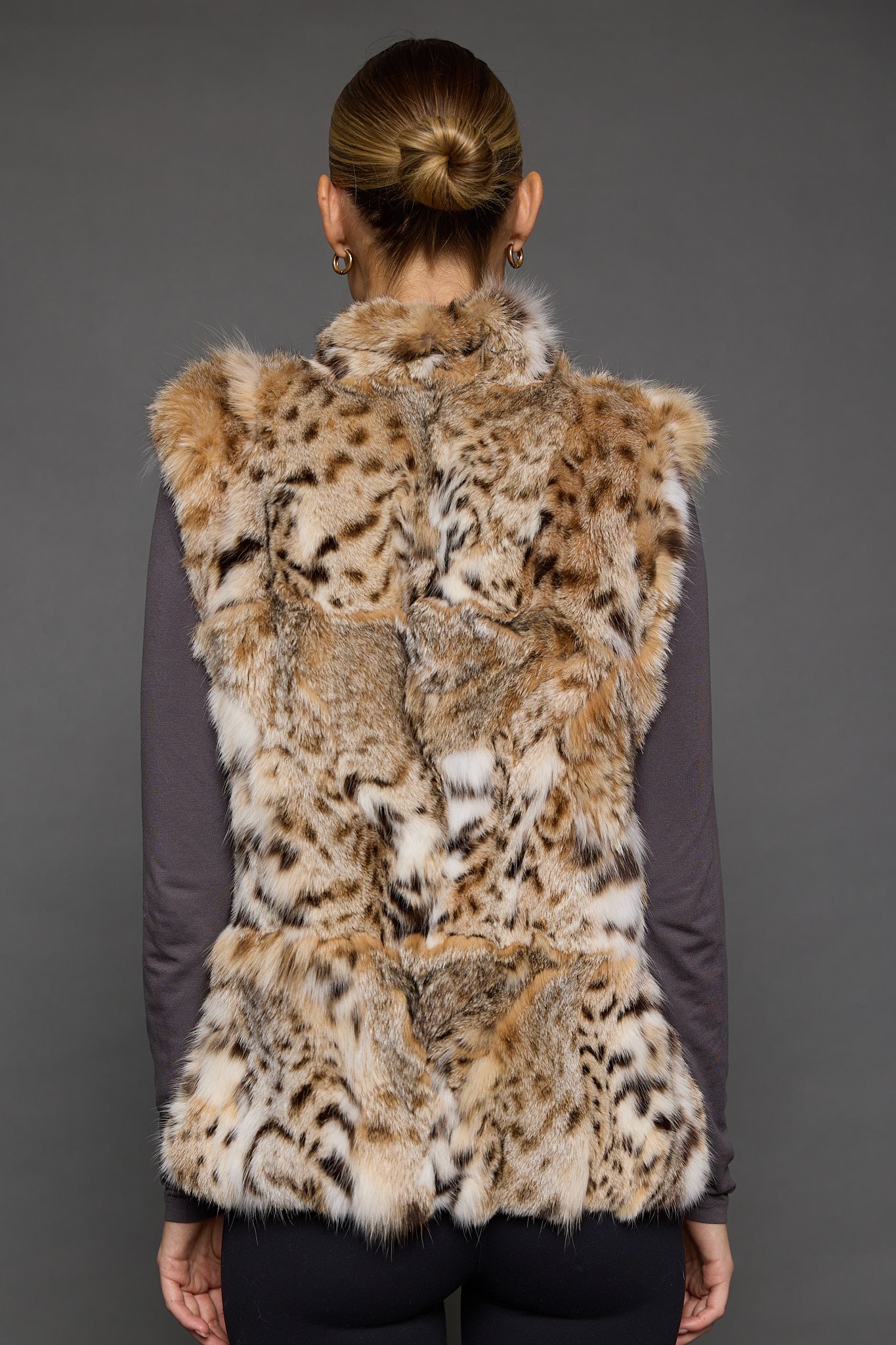 Rear view of a woman in a sculpted American lynx vest with her hands at her side, standing in front of a grey background