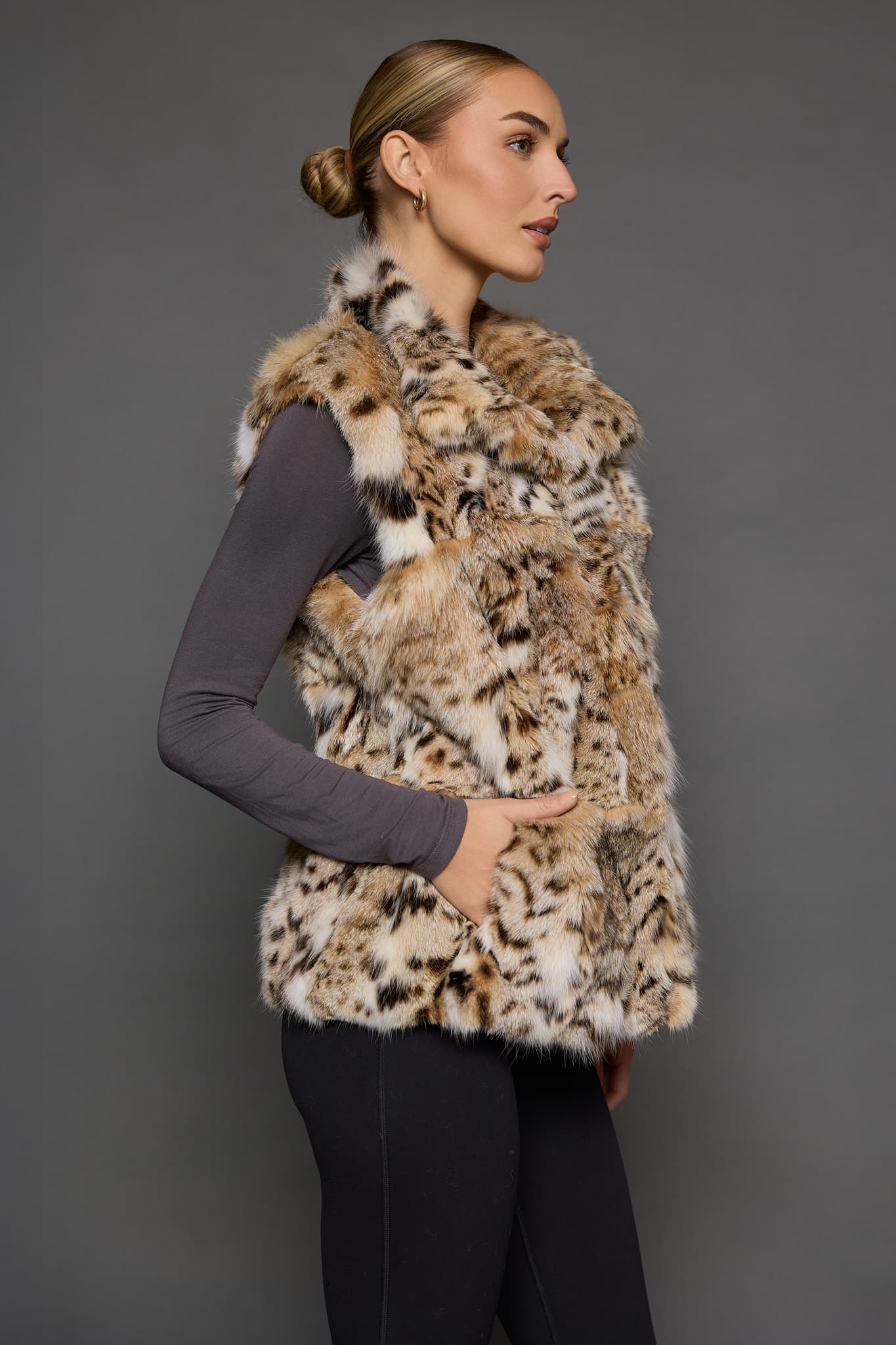 Side view of a woman in a sculpted American lynx vest with her right hand in the front pocket, standing in front of a grey background