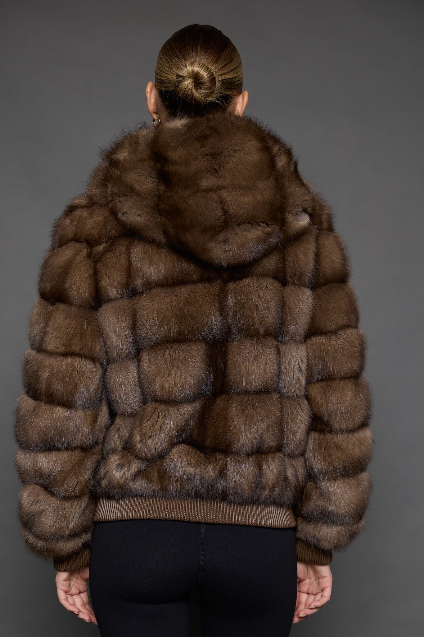 Rear view of a woman in a sable jacket with the hood down facing a dark grey background