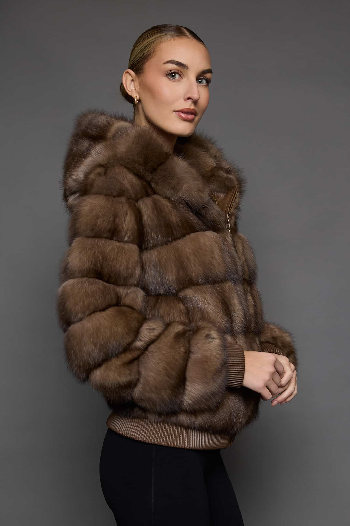 Profile view of a woman in a sable jacket with the hood down standing in front of a dark grey background
