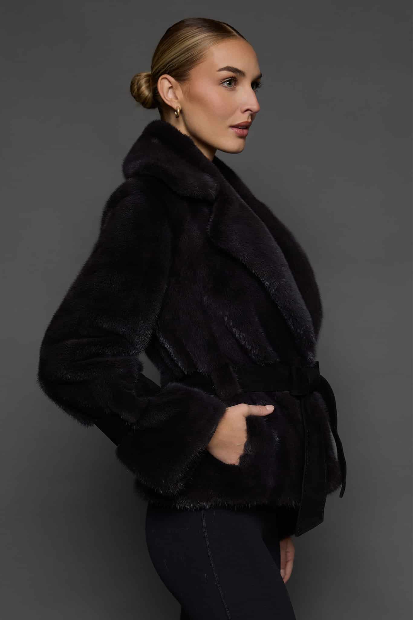 Profile shot of a woman in a sapphire mink jacket with her right hand in the pocket standing against a dark grey background