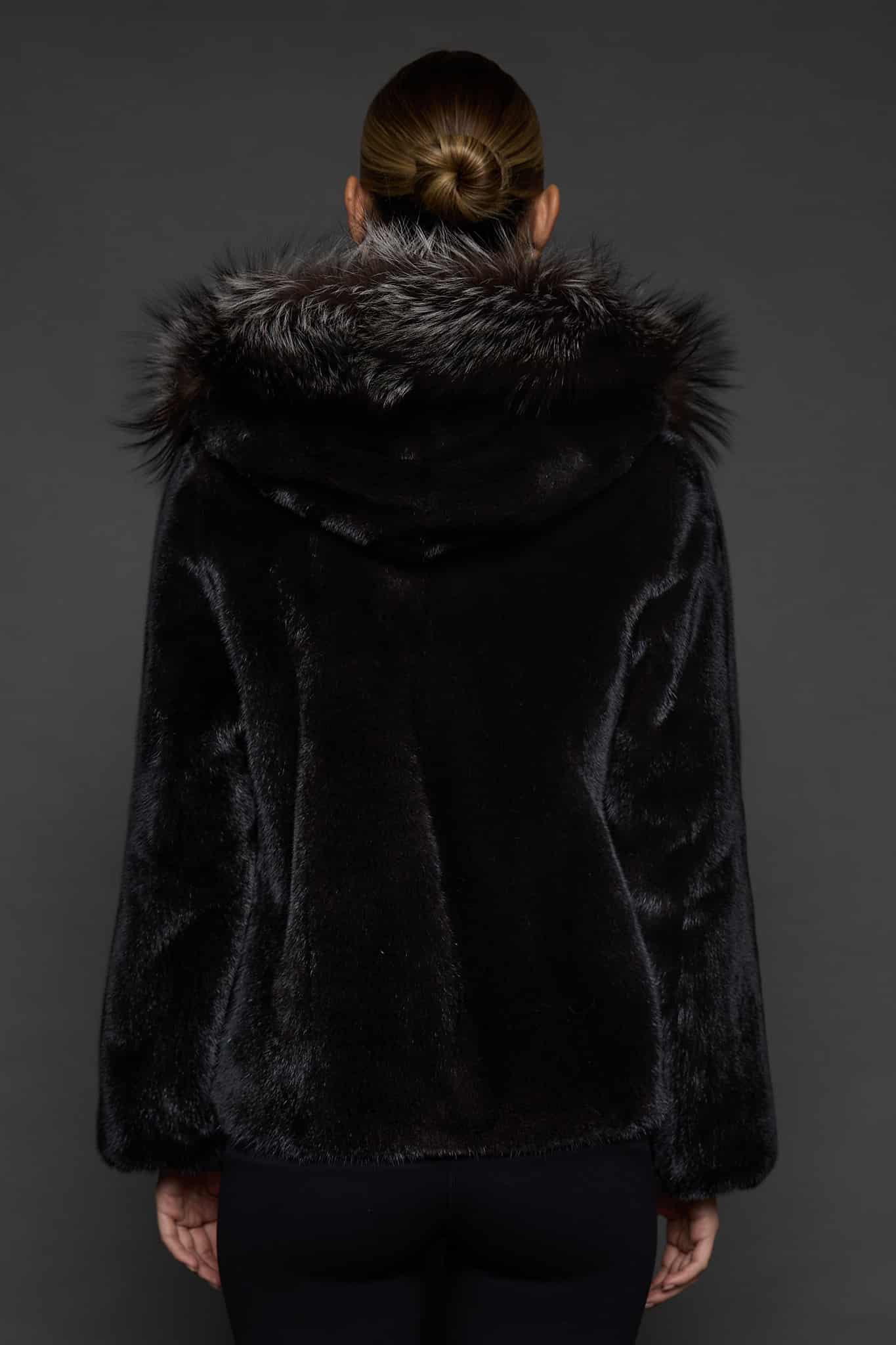 Rear view of a woman in a black mink jacket with the hood down standing in front of a dark grey background