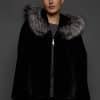 Frontal view of a woman in a black mink jacket with the hood down standing in front of a dark grey background