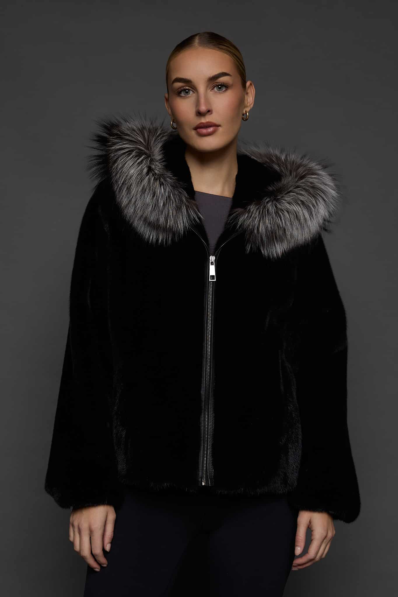 Frontal view of a woman in a black mink jacket with the hood down standing in front of a dark grey background
