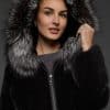 Close-up frontal view of a woman in a black mink jacket with the hood up standing in front of a dark grey background