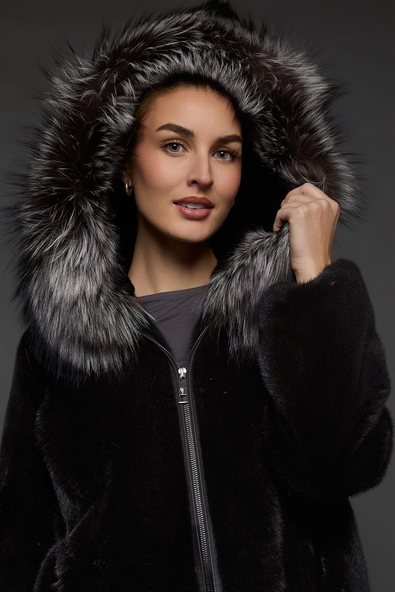 Close-up frontal view of a woman in a black mink jacket with the hood up standing in front of a dark grey background