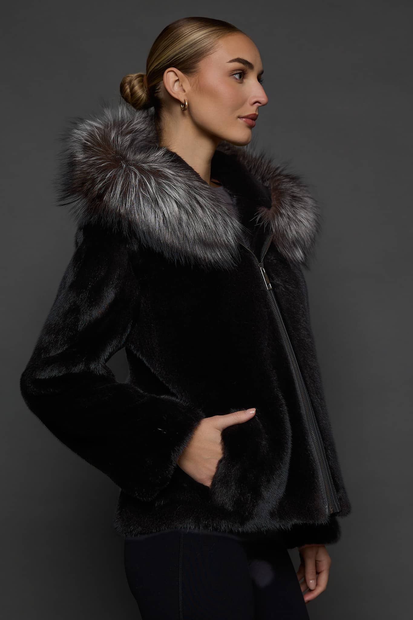 Profile view of a woman in a black mink jacket with the hood down standing in front of a dark grey background