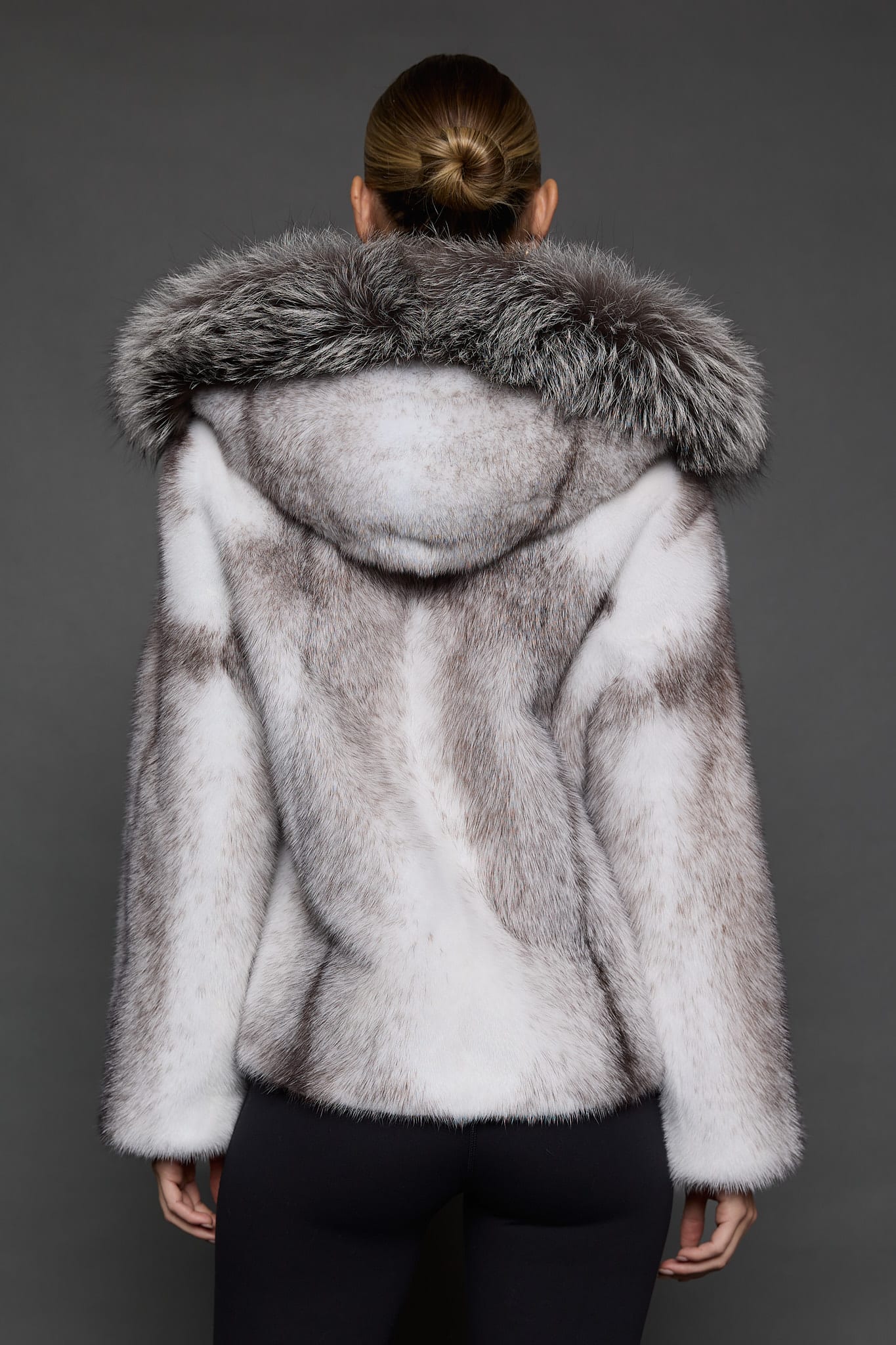 Rear view of a woman in a black cross mink jacket with the hood down facing a dark grey background